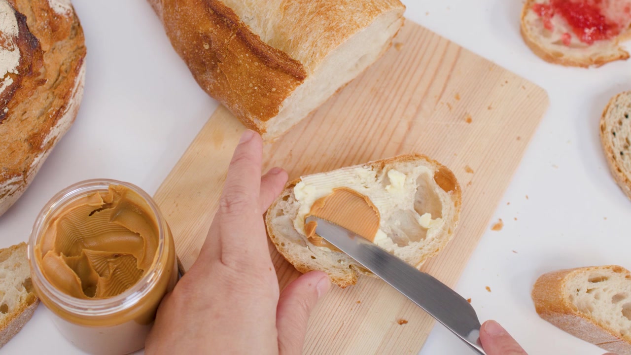 Mani usano un coltello per spalmare burro di arachidi cremoso su fette di pane a lievitazione naturale fresco