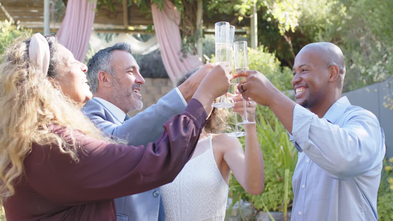 Diverse group lifting bubbly flutes, toasting couple after man inspecting glass under draped arch