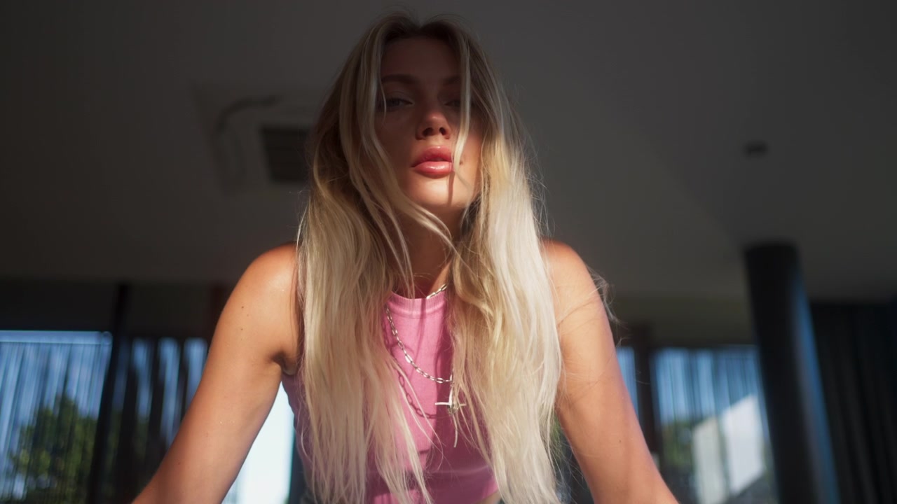 Primo piano al rallentatore di una bella donna bionda, con top rosa e jeans, seduta in un appartamento moderno e soleggiato, con un'espressione concentrata e contemplativa. Illuminazione naturale e soffusa, atmosfera di stile di vita urbano e domestico.