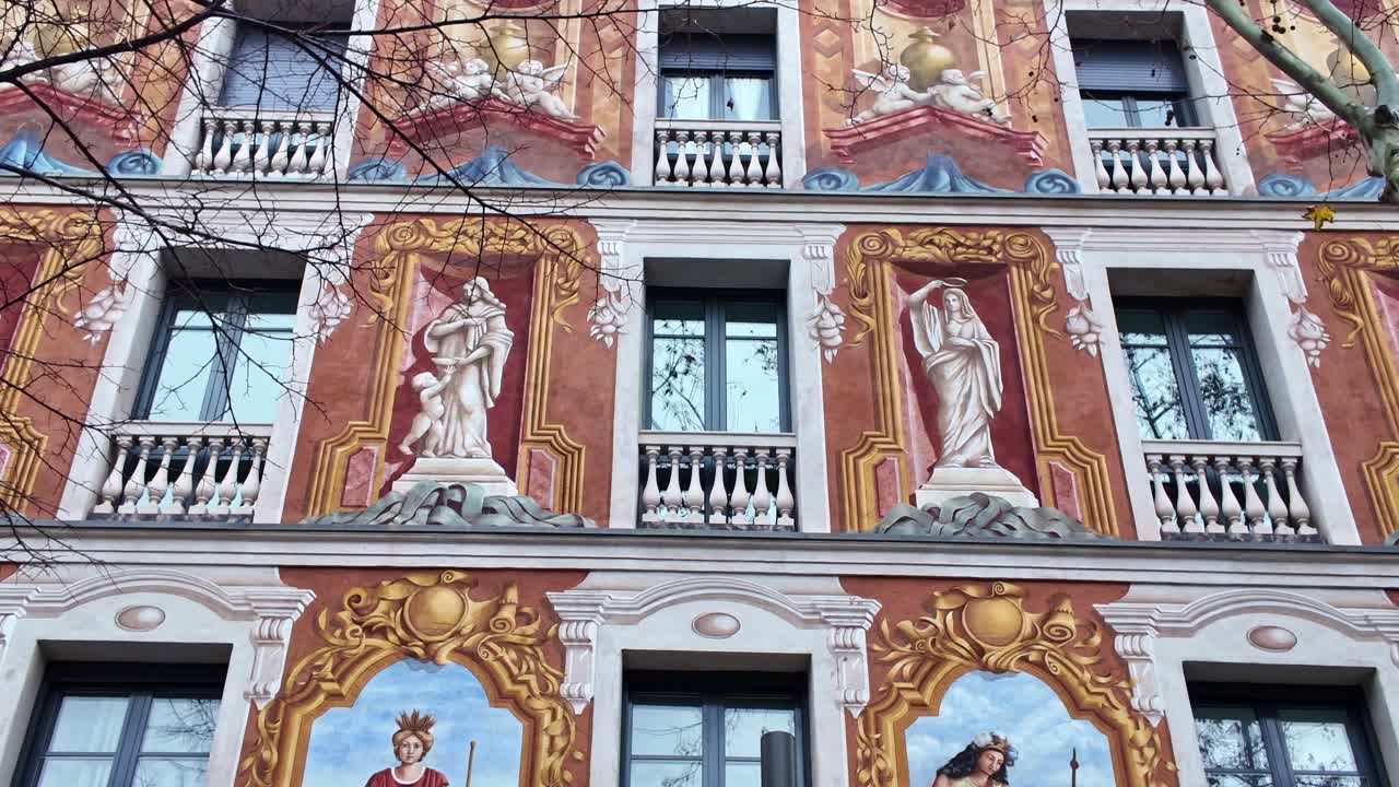 Inquadratura dal basso verso l'alto della facciata dipinta di un edificio decorativo a Barcellona, con figure allegoriche in stile classico e dettagli architettonici ornati. Spagna