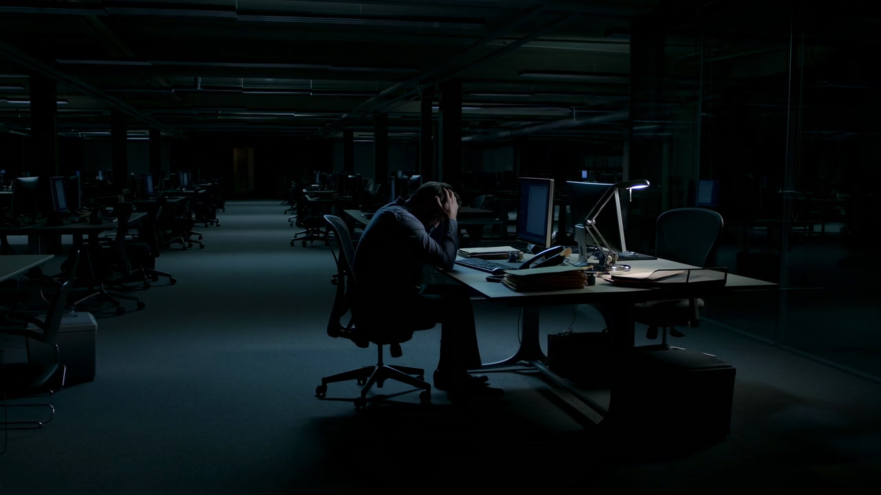 Late Night Office Worker Stressed At Desk, Illuminated By Single Monitor And Lamp, Hunched With Head In Hands