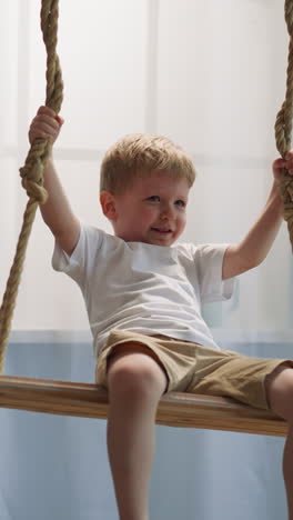 smiling toddler boy sits on swing suspended in living room. cute male kid wearing white t-shirt and light brown shorts rests at home low angle shot