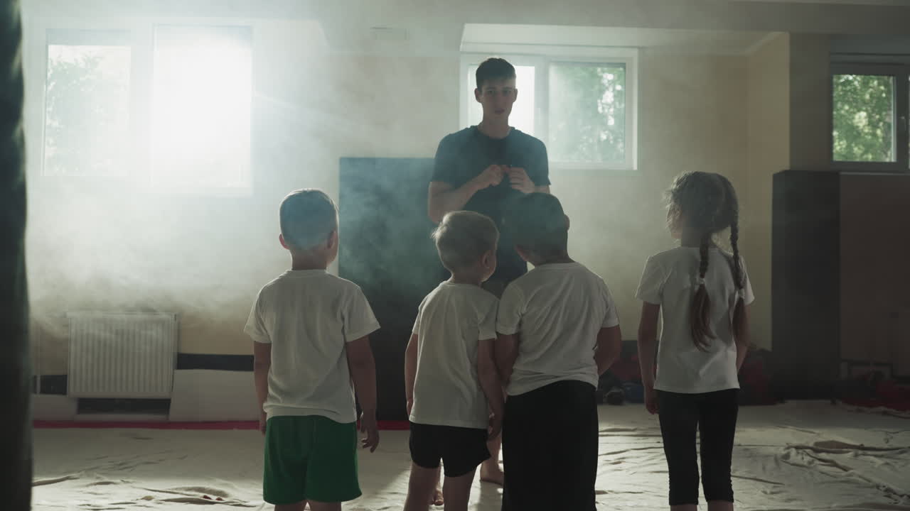 체육관에서 전투 수업에서 트레이너와 함께 작은 아이들. 귀여운 초등 학생들은 연기 클럽 느린 모션에서 싸우기 전에 강사를 듣습니다.