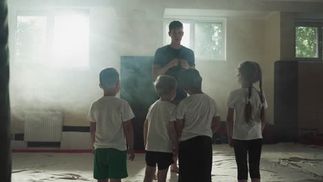 little children with trainer at combat lesson in gym. cute junior students listen to instructor before fighting in smoky club slow motion. martial arts