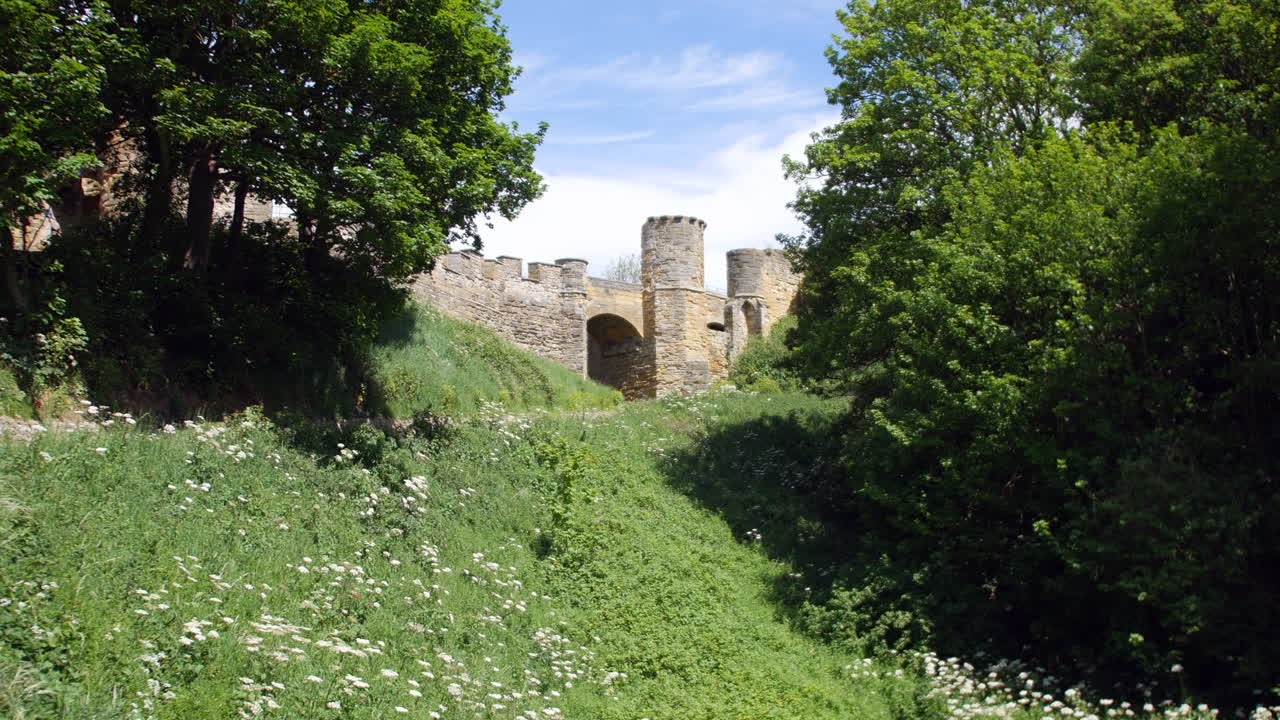 Slotsruiner står over en græsklædt skråning i Scarborough, North Yorkshire, England, indrammet af træer og vilde blomster med middelalderlige stenmure og tårne.