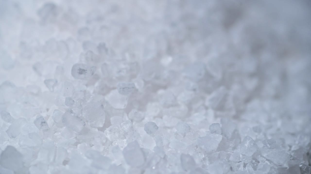 An extreme close-up macro shot of coarse sea salt crystals. The irregular shapes and translucent texture of the salt grains are highlighted, creating a detailed and natural composition.