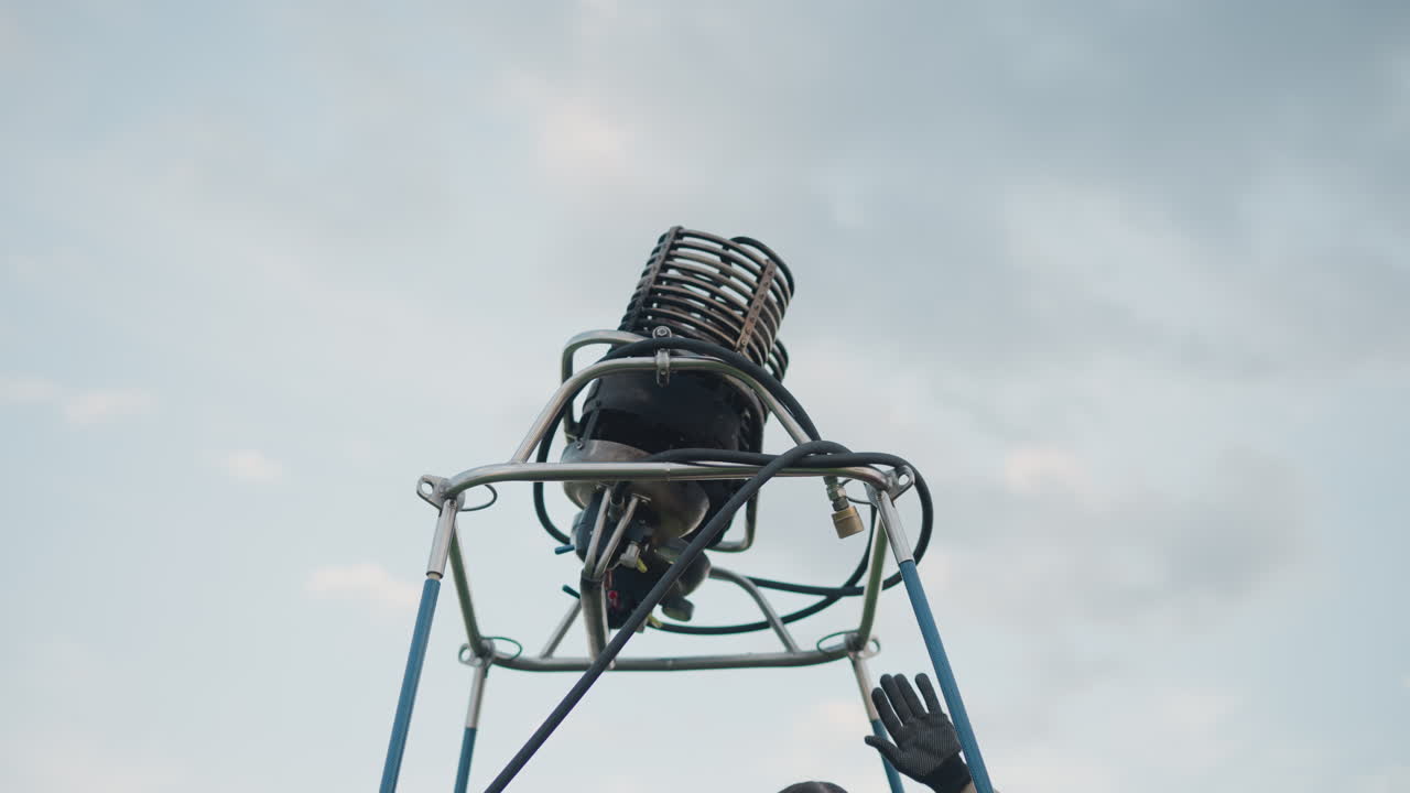 woman adjusting metal burner on hot air balloon frame poles stabilizing wobbling component under cloudy sky roadside field while partner holds support rods during preflight setup before flight launch