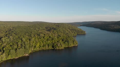 slow aerial view of lake in quebec surrounded by trees at sunrise with the sun shining over the hill and trees