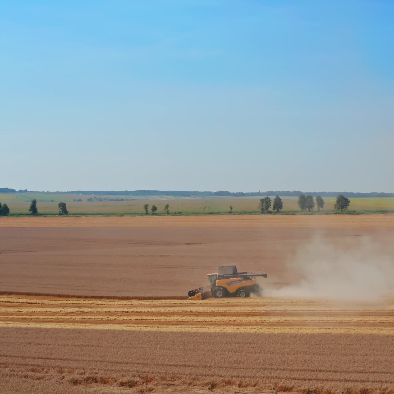 Beautiful countryside scenery with agribusiness plantations. Yellow harvester working in the field at foreground. Blue clear sky backdrop