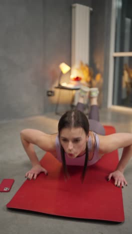 close-up vertical video of a confident brunette girl in a sports summer uniform doing push-ups and sprints on a red sports mat in a sports mat in a modern apartment at home