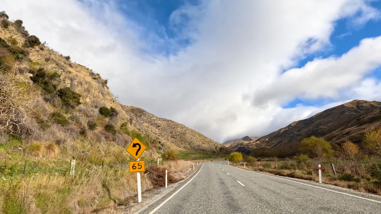 A car travels through Wanaka's picturesque mountain roads under a bright, partly cloudy sky, showcasing stunning natural scenery