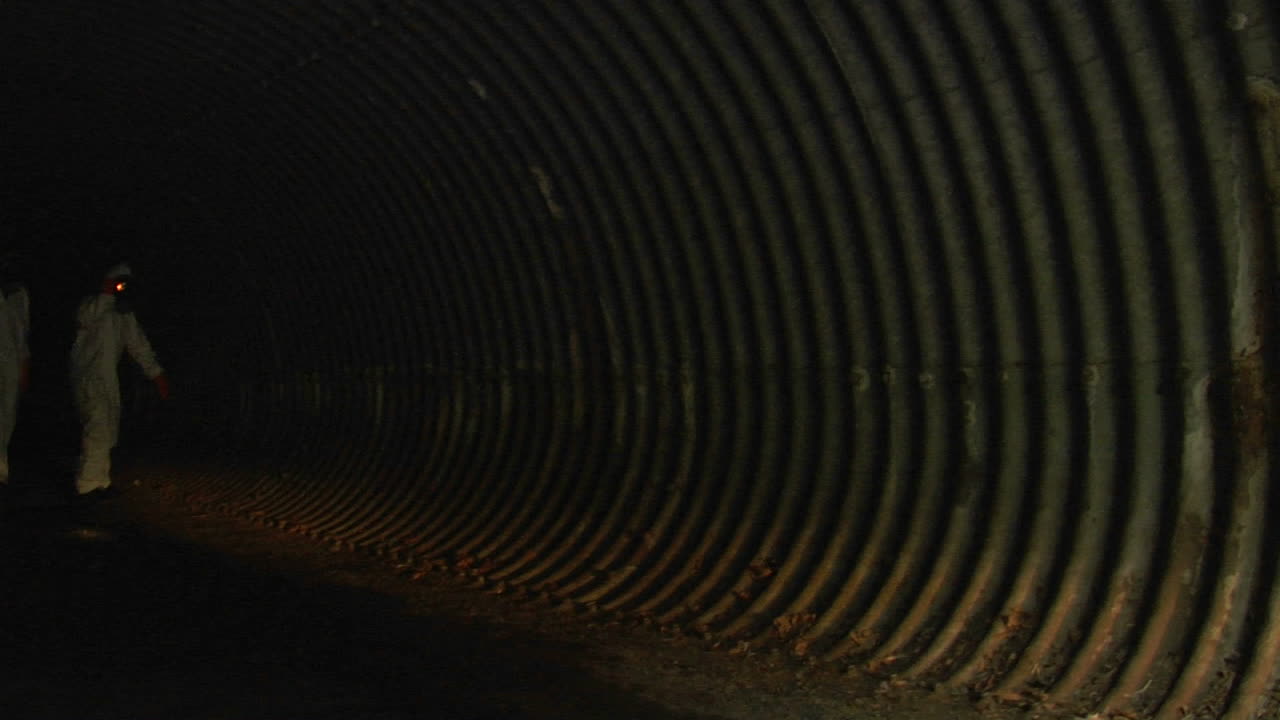 방호복을 입은 개인이 손전등을 사용하여 어두운 터널을 조사합니다.