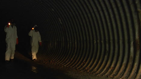 individuals wearing hazmat suits use flashlights to inspect a dark tunnel