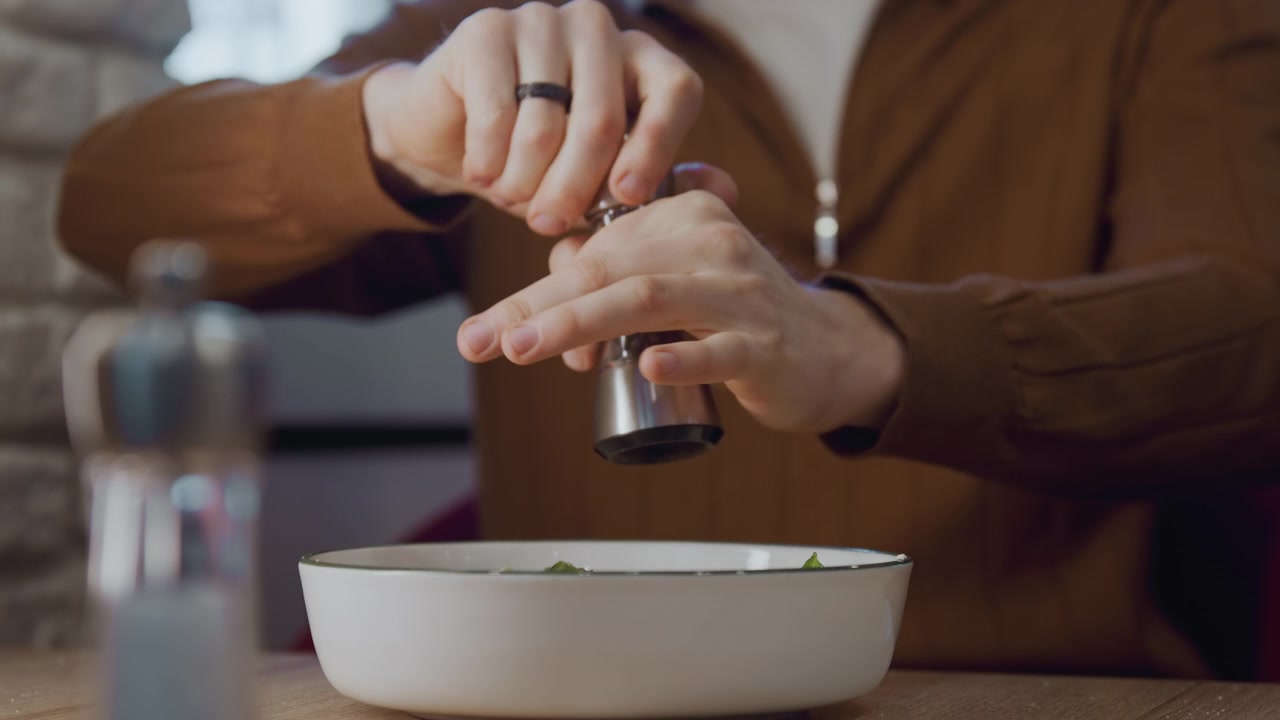 Primo piano di una persona con abiti marroni che macina il pepe in una ciotola bianca con erbe aromatiche verdi. Le mani azionano un macinino di metallo sopra un piatto poco profondo, concentrandosi sulla preparazione del condimento. Illuminazione interna soffusa