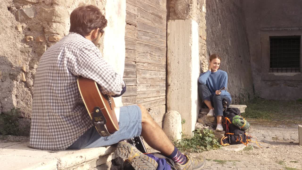 pareja feliz de mochileros turistas tocando la guitarra y cantando sentados cerca de la puerta de madera de un edificio medieval en el parco degli acquedotti ruinas del parque en roma al amanecer con guía de mapa cámara constante en cámara lenta
