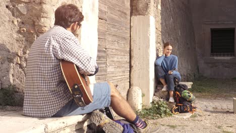 happy young couple backpackers tourists playing guitar and singing sitting near wooden doorway of medieval building in parco degli acquedotti park ruins in rome at sunrise with map guide slow motion steadycam