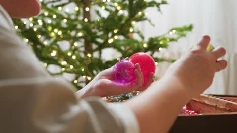 hand view of woman picking colorful ornament balls from box beside decorated christmas tree, preparing festive decorations with care and joy in cozy home environment during holiday season