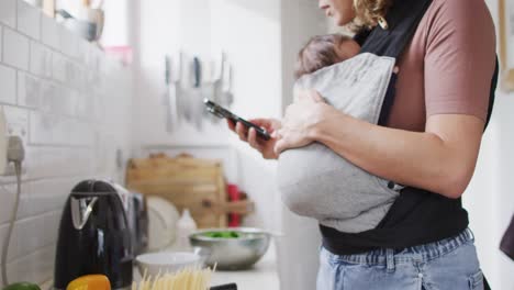 Video-De-Una-Madre-Caucásica-Con-Un-Bebé-Recién-Nacido-En-Un-Portabebés-Usando-Un-Teléfono-Inteligente-En-La-Cocina