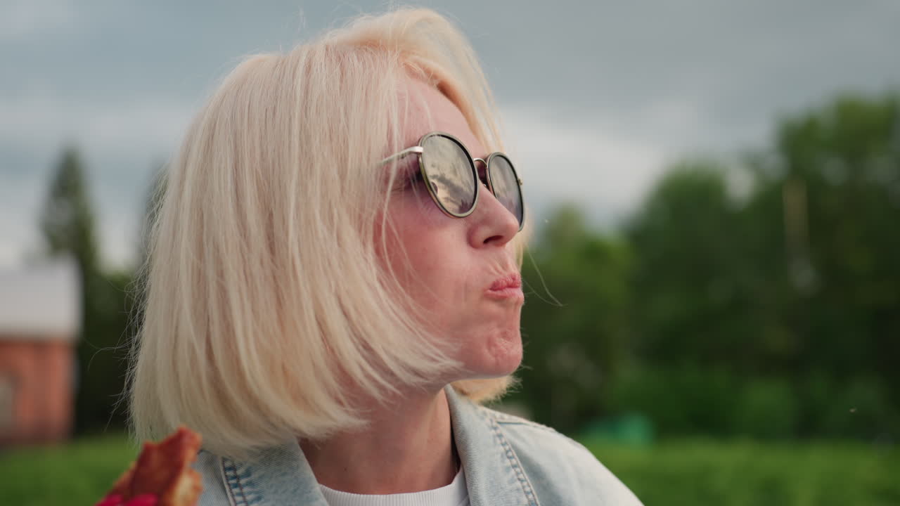 Una donna elegante con occhiali da sole addenta una fetta di pizza all'aperto, profilo laterale che mostra mentre assapora il condimento al formaggio con la luce del tramonto e un'espressione rilassata in un parco verde.