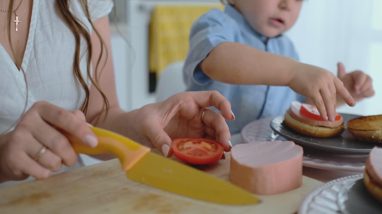 una madre joven con un niño pequeño juntos cortados con un cuchillo un tomate para una hamburguesa casera. comida saludable cocinar juntos