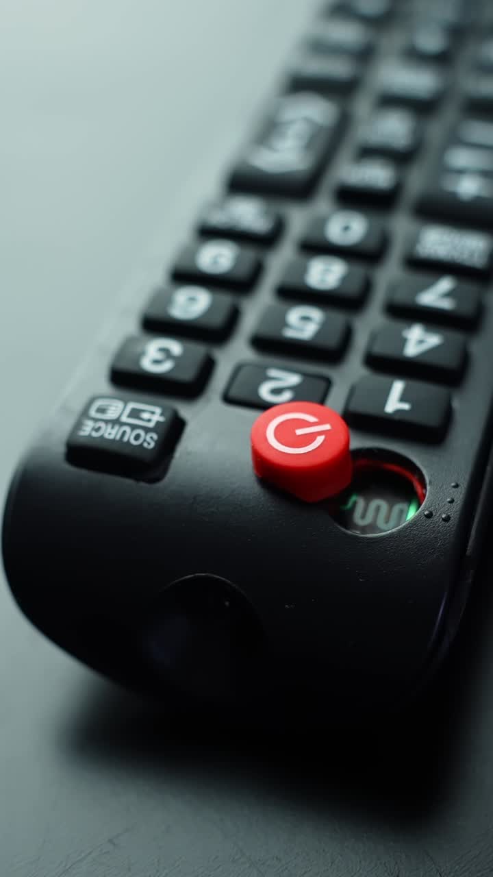 Inquadratura verticale con messa a fuoco selettiva, da un macro nitido su un pulsante di accensione rosso a una visione più ampia e sfocata di un telecomando TV su sfondo grigio con spazio per il testo.