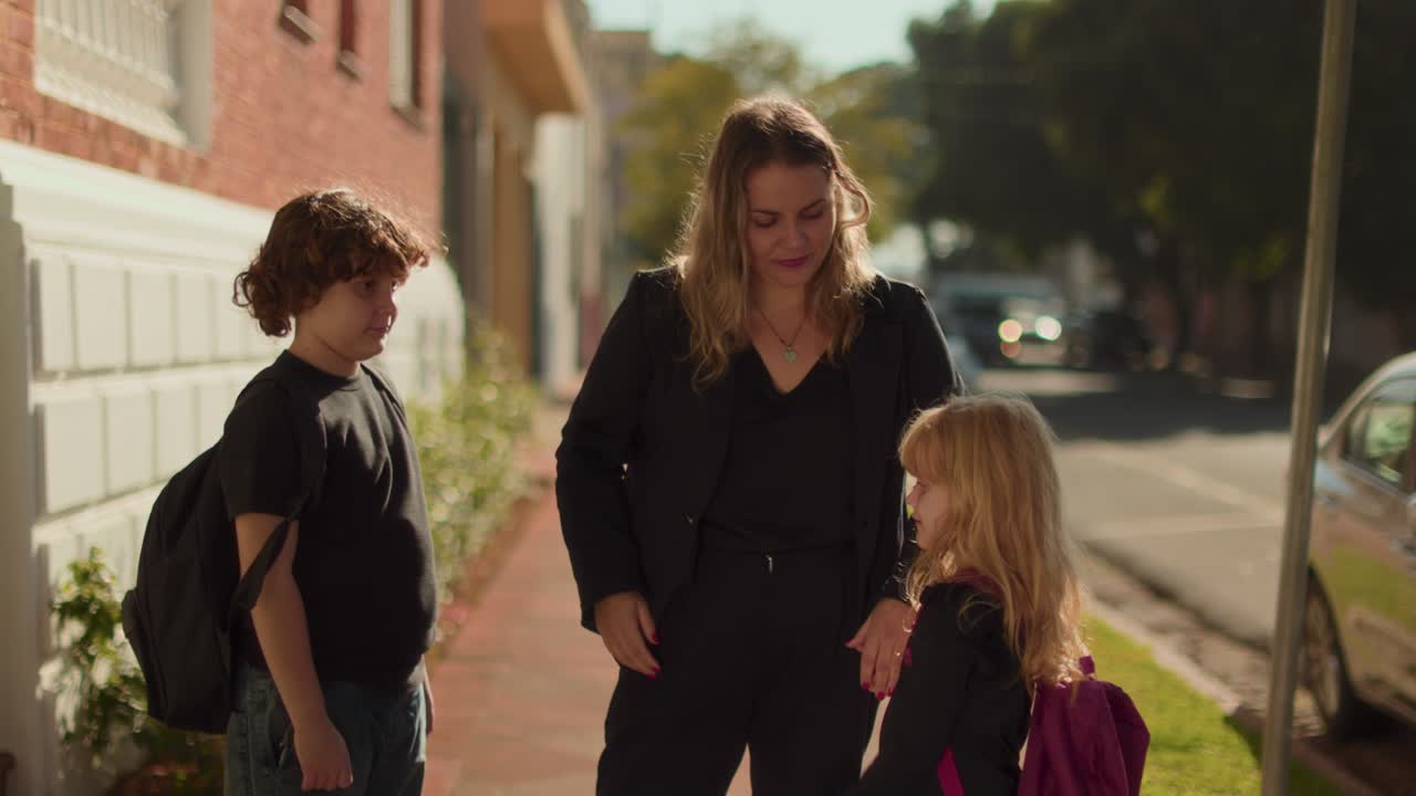 A mother sends her two children, a son and a daughter, off to school with goodbyes and affectionate gestures on a sunny urban sidewalk