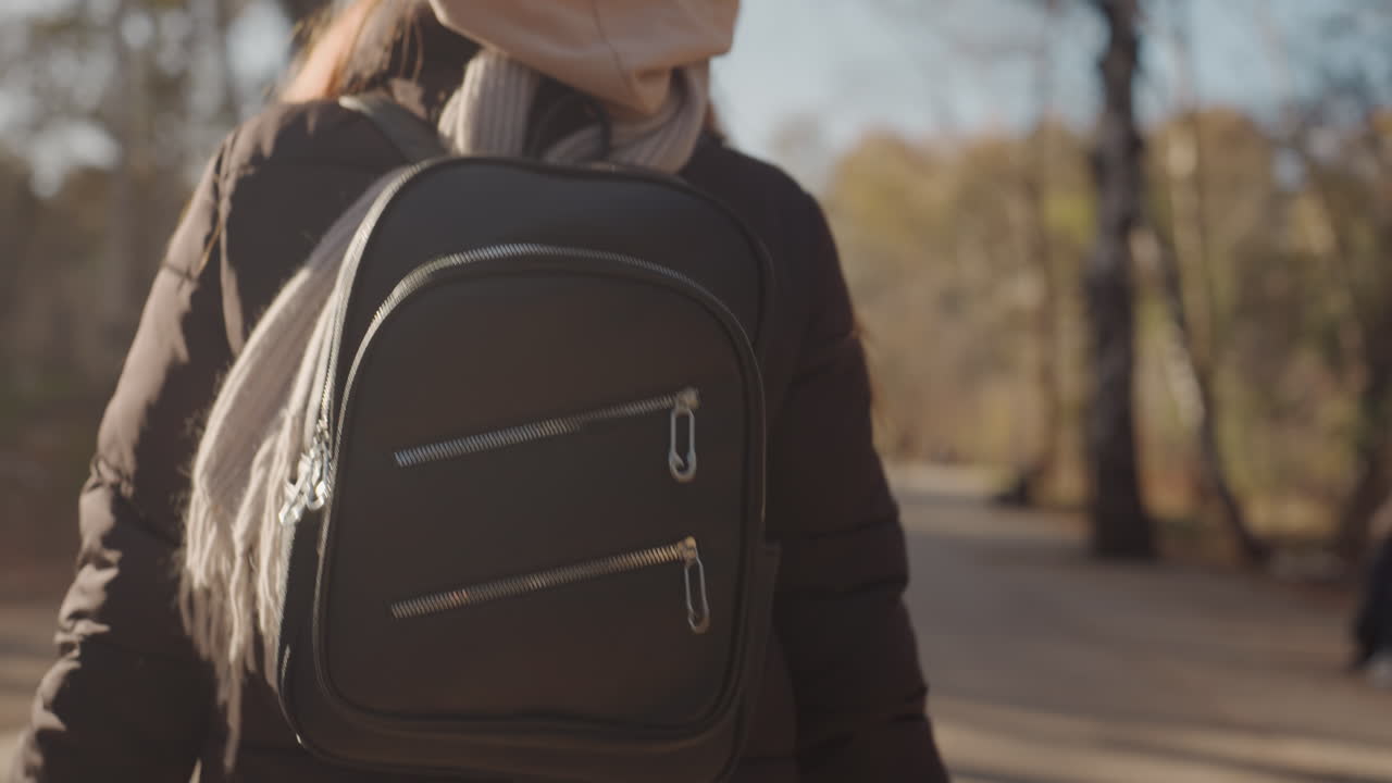 Woman with backpack walking in park. Autumn afternoon, puffer jacket and scarf, closeup on bag and straps, subtle tension suggests possible snatcher nearby, cinematic warm tones and candid street