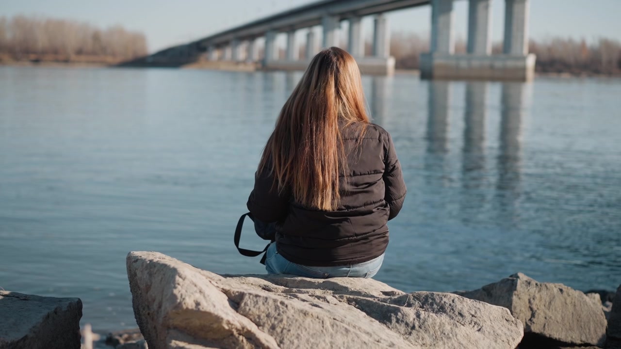Donna seduta su una roccia vicino all'acqua, di spalle alla fotocamera, con capelli lunghi e abbigliamento casual. Lo sfondo presenta un fiume calmo con riflessi di alberi e una struttura di ponte sopraelevato sotto una luce soffusa.