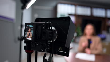 caucasian woman wearing beige jacket speak on the camera display. blogger sits at desk at blurred backdrop talking to camera.