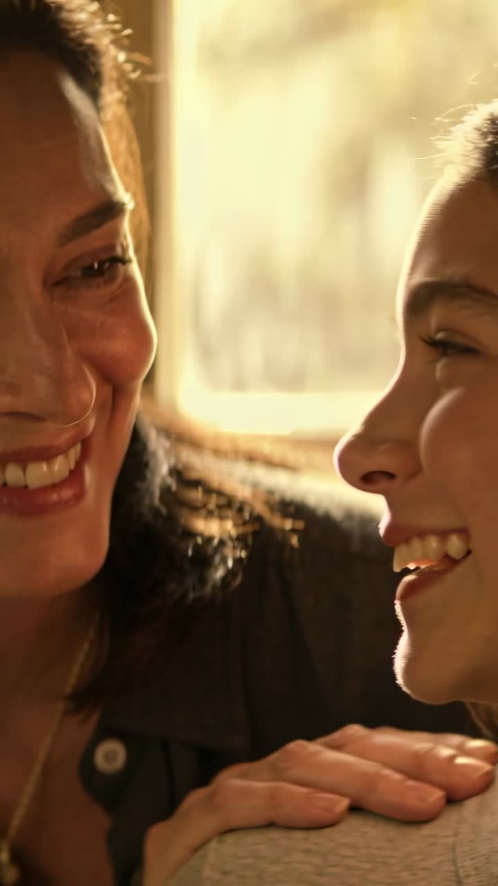 Vertical video: Zooming camera on daughter, mom leaning, hand on shoulder at window, sharing smile