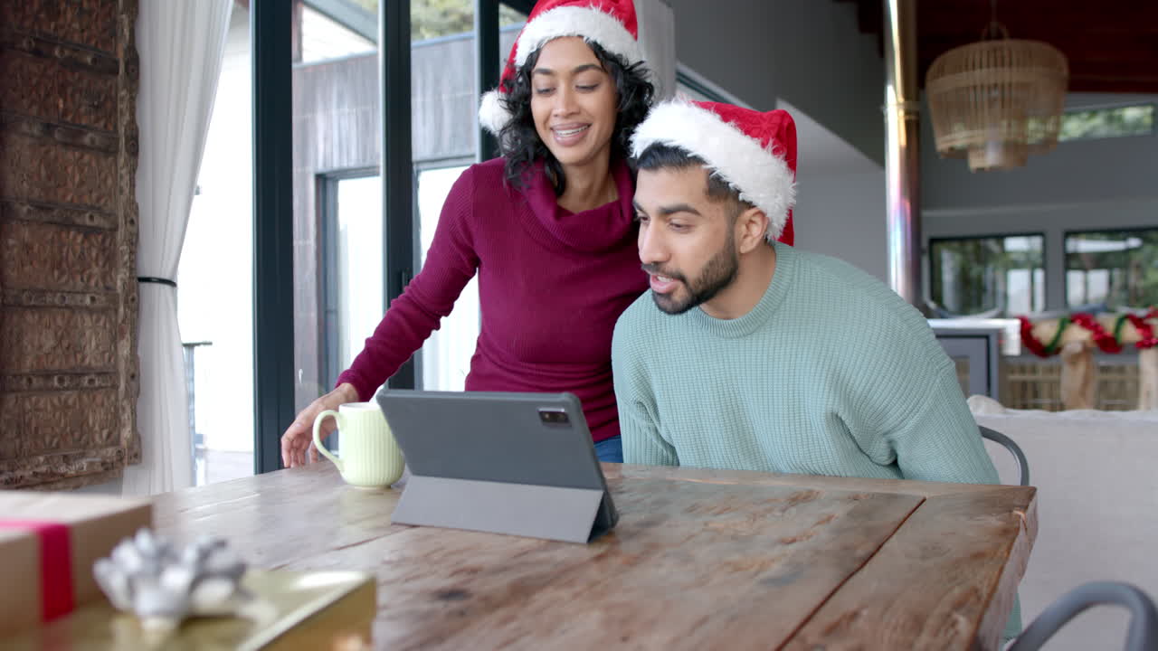una feliz pareja biracial con sombreros de papá noel usando una tableta para una llamada de video en casa, en cámara lenta