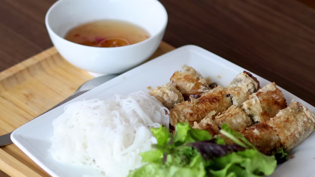 Close-up of spring rolls, rice noodles, and fresh greens on a white plate with dipping sauce.