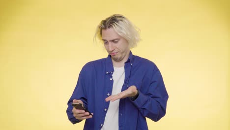 A-young-man-in-a-blue-shirt-points-at-his-smartphone-and-disappointed-with-what-he-sees,-yellow-background