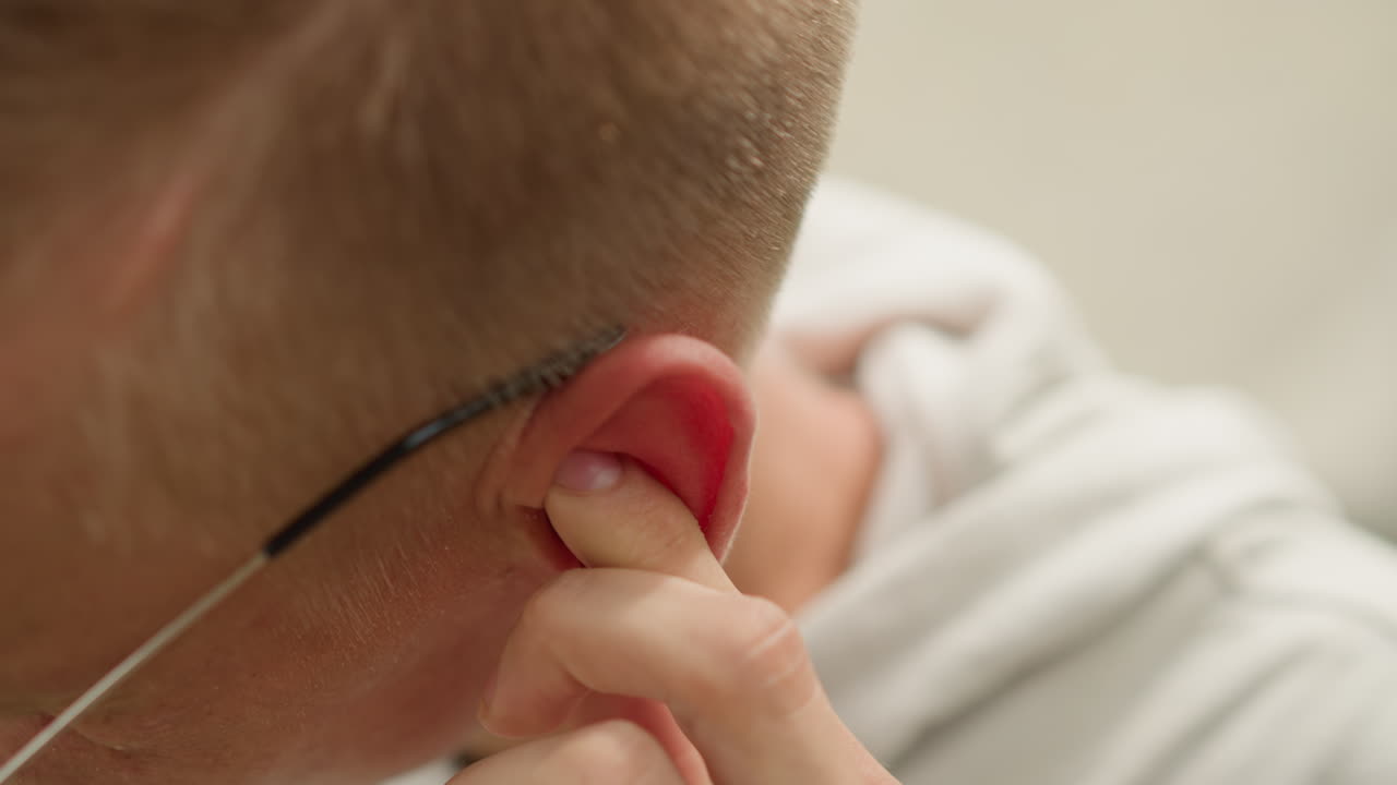 Primer plano de un hombre blanco tocándose el oído interno, con detalle de la yema del dedo y el enrojecimiento de la oreja; la secuencia sugiere a un músico afinando, a un corredor quitándose agua y a un estudiante comprobando si tiene alguna irritación, encuadre íntimo en interior, suave y neutro