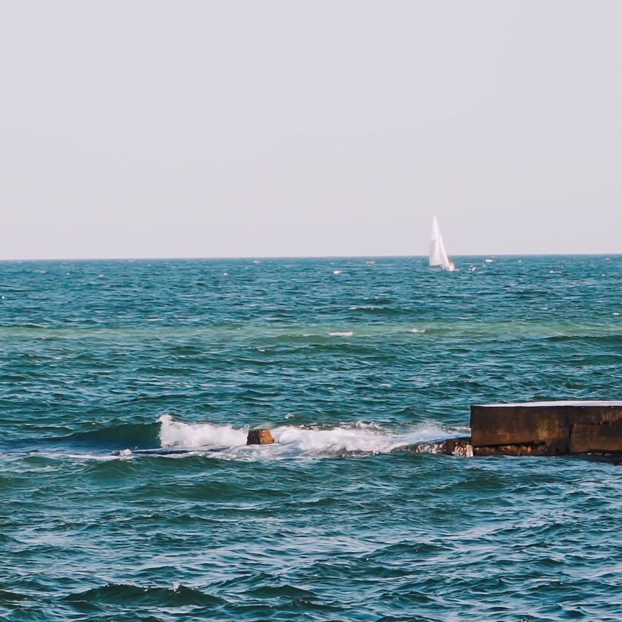 Motion of sea water. Waves crashing on stone harbor making white foam. White sail floating on the water surface. Slow motion.