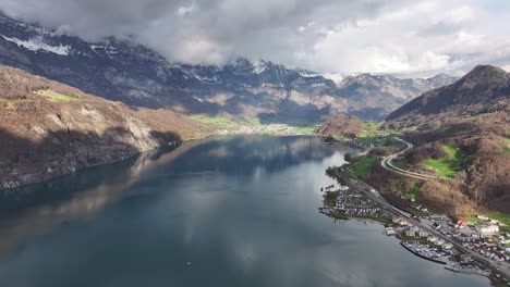 Vista-Aérea-Desde-Gran-Altura-Sobre-Walensee,-Suiza,-Mostrando-Un-Telón-De-Fondo-De-Montañas-Cubiertas-De-Nieve-Y-Una-Pintoresca-Zona-Residencial-Ubicada-A-Lo-Largo-De-Las-Orillas-De-La-Costa.