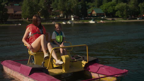 joyful woman and schoolgirl talk sitting in catamaran. relaxed mother shows daughter boat passing by slow motion. sight-seeing at resort with reservoir