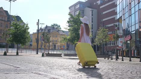 a slim beautiful woman in a white dress and hat walks with a yellow suitcase in the old city. a female tourist is heading to the hotel. summer vacation in europe