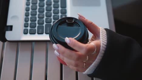 primer plano de la mano de la mujer con las uñas cuidadosas girando suavemente la taza de café cerca de la computadora portátil, centrándose en los dedos contra un fondo borroso del teclado