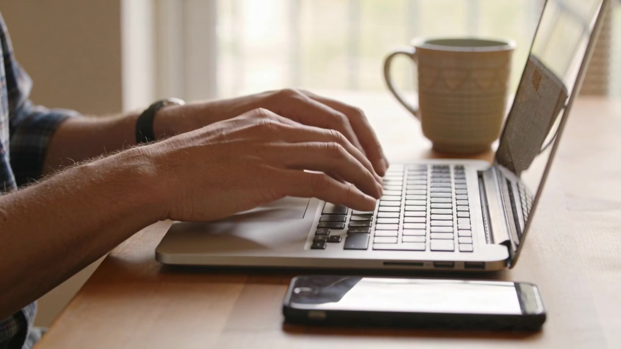 Inizio del lavoro, uomo con camicia a quadri e orologio che digita vicino alla finestra, che prende il telefono mentre è al laptop.