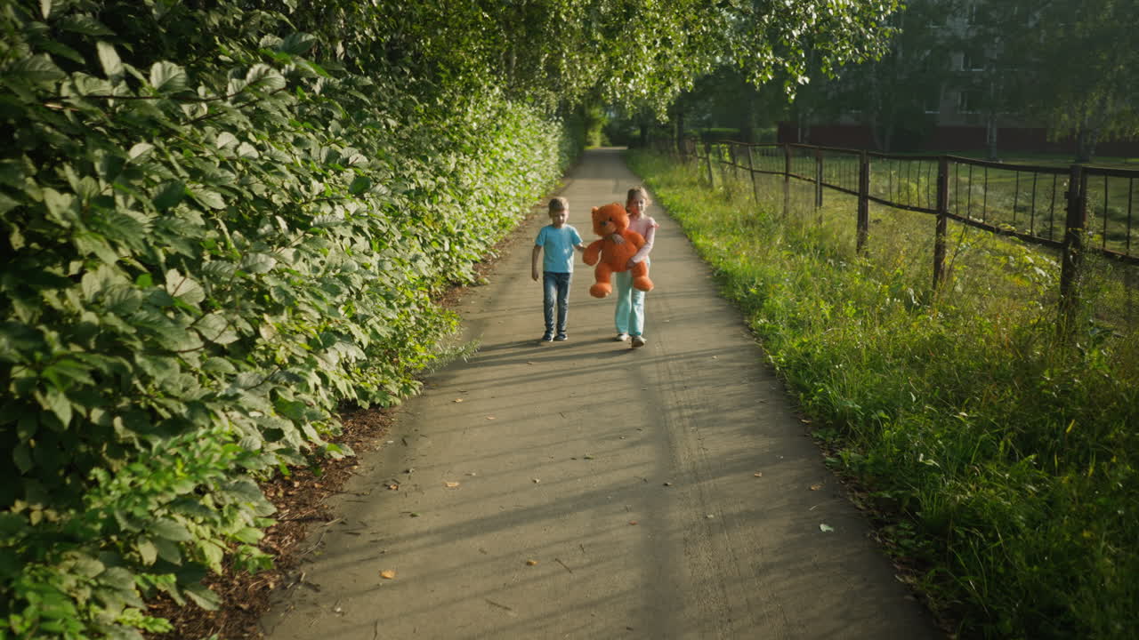 Girl holding teddy bear tightly while boy supports by hand as they walk along quiet tree lined path, warm sunlight casts long shadows across walkway bordered by tall greenery and iron fencing