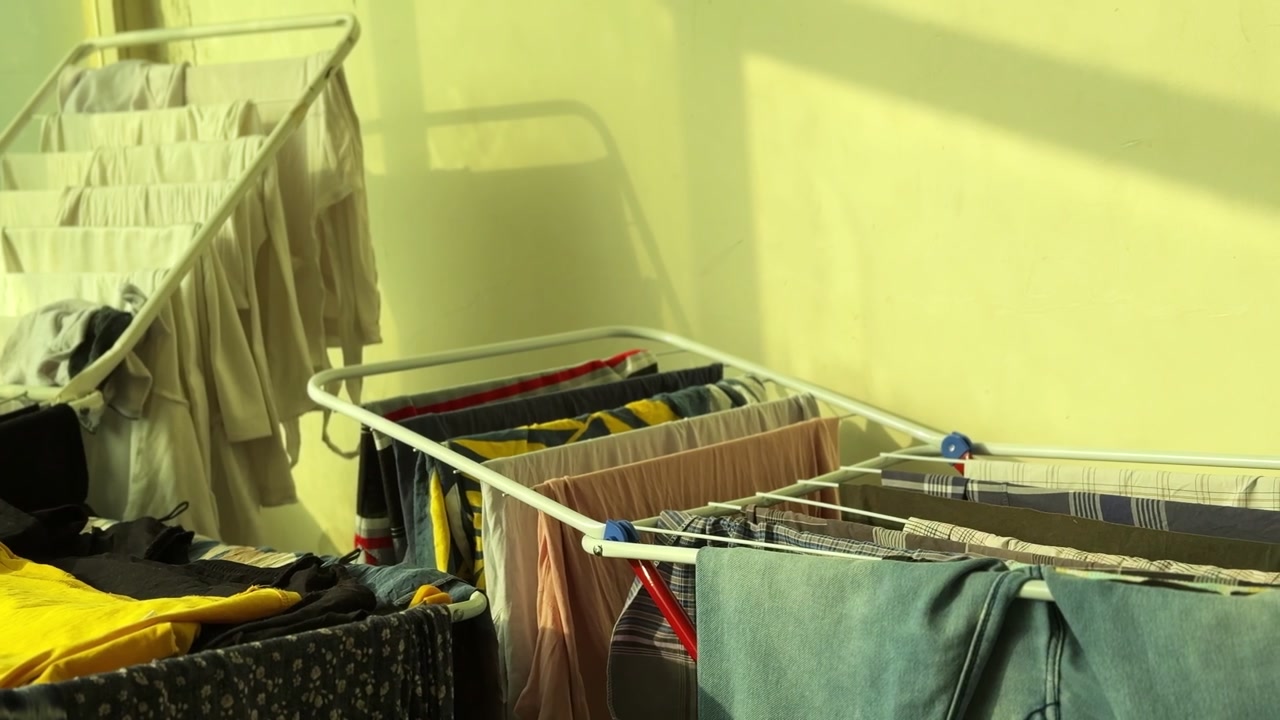 Multiple clothes racks filled with freshly washed garments dry indoors under warm sunlight, casting strong shadows across a yellow wall in a bright home laundry space.