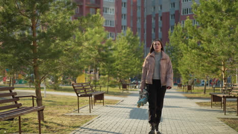 front shot of a girl walking through a sunlit park, wearing a peach jacket, black trousers, and black shoes. she is holding a pair of rollerblades, with trees and benches lining the pathway