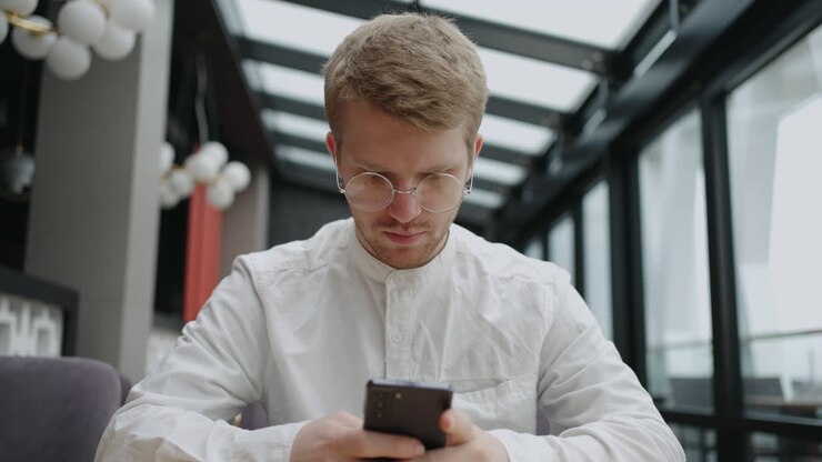 handsome caucasian man with eyeglasses is using smartphone for chatting and sending email sitting alone in cafe
