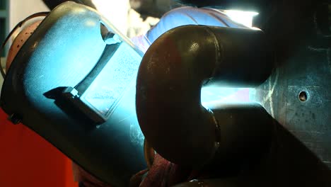 reverse shot of a welder in full protective hse kit and face mask welding pipes at a factory with intense light and sparks from the welding operation