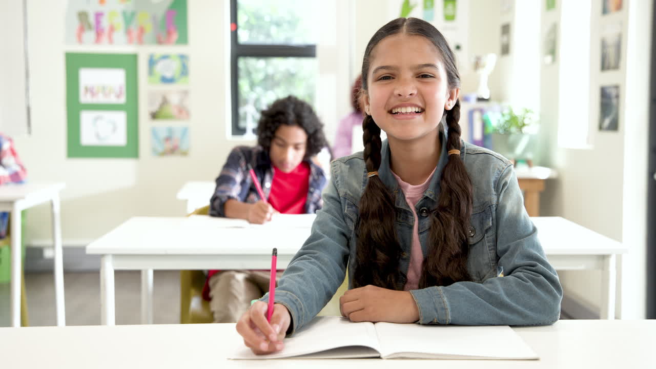 Smiling girl in school classroom writing in notebook, enjoying learning with multiracial students