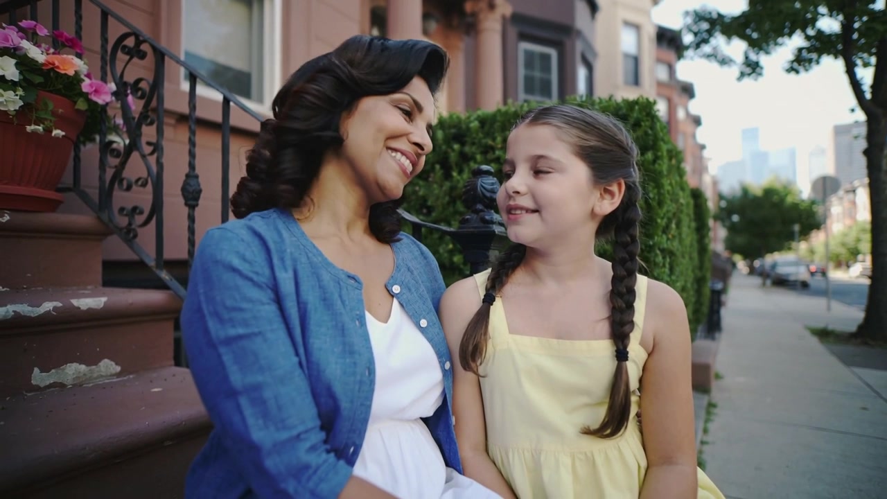 Mamá sonriente inclinándose hacia la niña que habla en el porche, con cárdigan azul y vestido amarillo, junto a una maceta, en un momento de unión