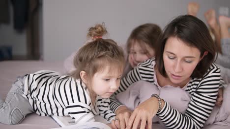 Young-mother-is-reading-a-tale-for-her-lovely-little-daughters-lying-on-a-bed.-Little-one-is-looking-through-the-book.-Close-up