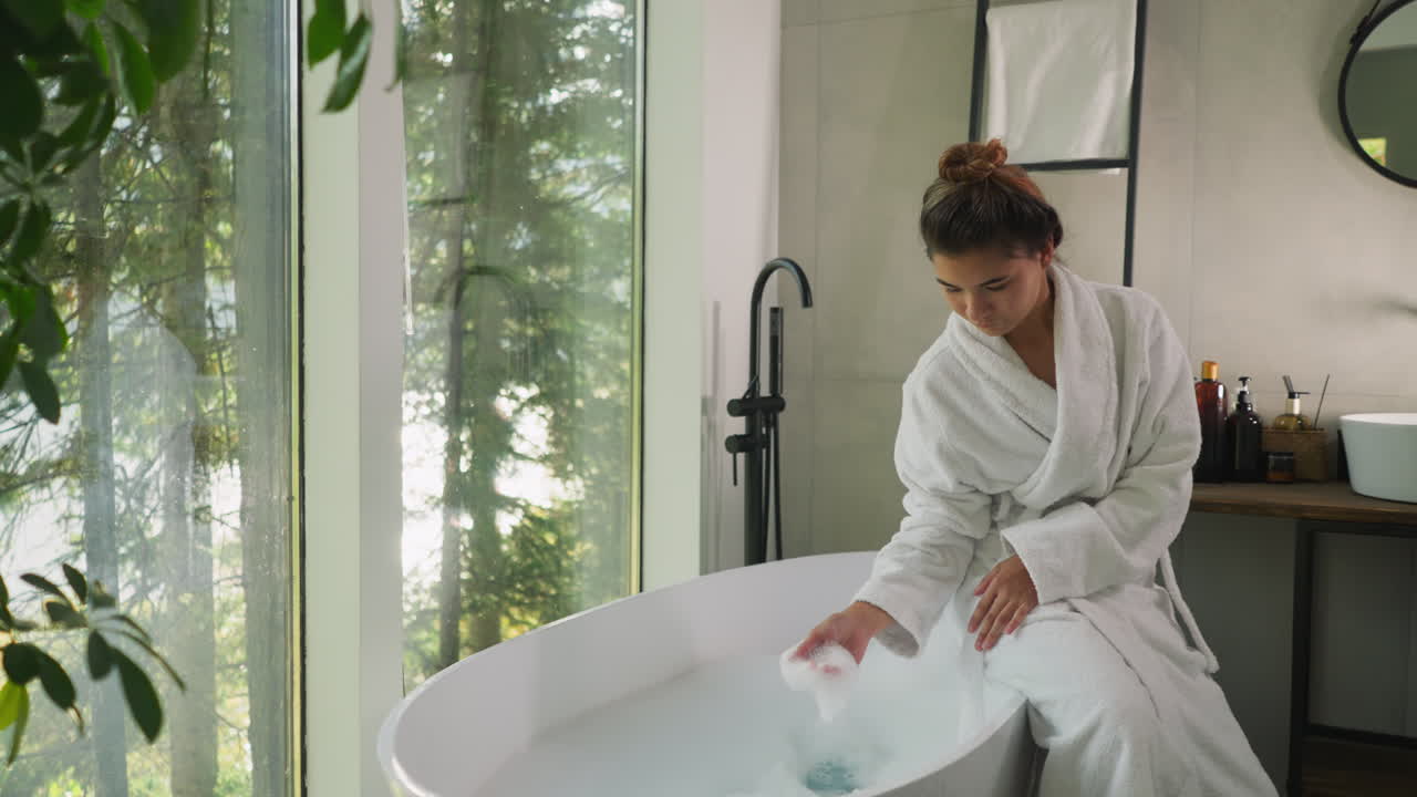 욕조 가장자리에 앉아있는 여자가 거품을 만지며, 테리 가운을 입은 매력적인 젊은 여자가 우아한 욕실에서 부드러운 거품으로 놀고 있습니다.