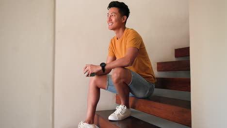 smiling young man sitting on wooden stairs at home, holding smartphone, copy space